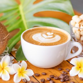 Latte in a cup with latte art, surrounded by kona coffee beans and plumeria flowers