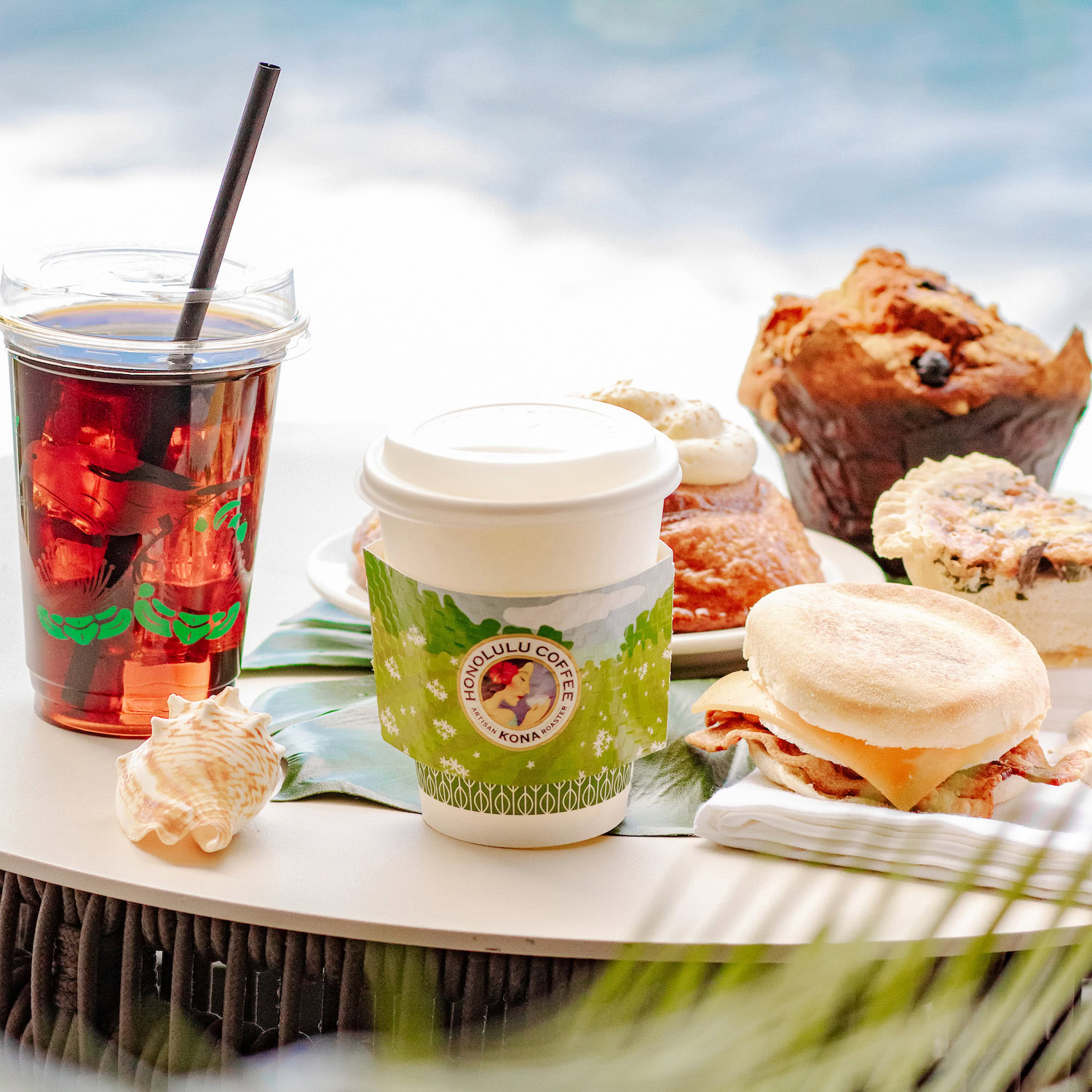 iced and hot coffee drinks from Honolulu Coffee on a table by the ocean with some sweet pastries on the side